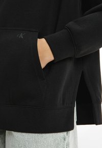Felpa nera in cotone con fessura laterale, polsini a coste, tasca frontale e un sottile accento con logo vicino all'apertura della tasca.