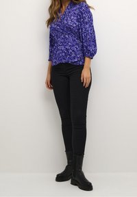 Blouse fleurie violette à manches trois-quarts, associée à un jean skinny noir et des bottines noires. Tissu lisse avec un design cache-cœur.