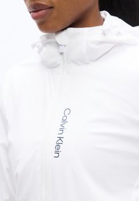 Κοντινό πλάνο ενός ατόμου που φοράει λευκό μπουφάν με φερμουάρ και κουκούλα, με το όνομα «Calvin Klein» τυπωμένο κάθετα στο στήθος.