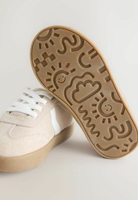 Beige suede sneakers with white accents and six white laces. The rubber sole features a textured pattern with playful designs and smiley faces.