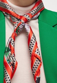 Sciarpa di seta rossa e a motivo legato attorno al collo, caratterizzata da disegni geometrici e una texture lucida, abbinata a un blazer verde.