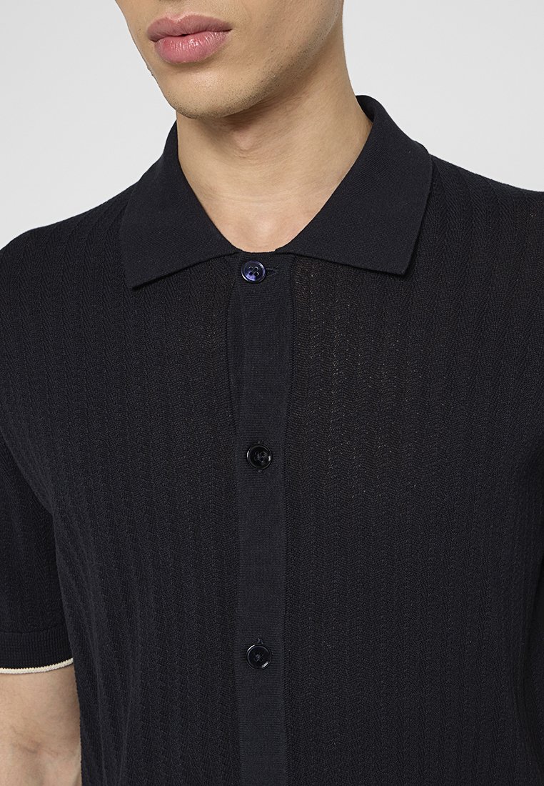 Ciemna, prążkowana koszulka polo z teksturowanej tkaniny, z czarnym zapięciem na guziki i kontrastującym jasnym wykończeniem przy rękawach.