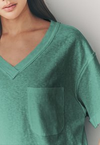 Tričko s výstrihom do "V" a krátkymi rukávmi vyrobené z textúrovanej tyrkysovej látky, so spodným vreckom na hrudi, ktoré zdôrazňuje voľný strih a jemné farebné odtiene.