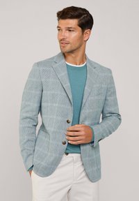 Blazer a quadretti blu chiaro in un tessuto strutturato con revers, e bottoni marroni, indossato sopra una maglia a maglia teal e pantaloni bianchi.