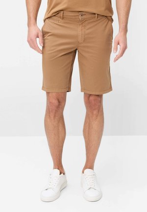 Mann trägt beige knielange Shorts und weiße Sneakers und steht vor einem schlichten weißen Hintergrund.