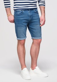 Mann trägt aufgekrempelte Jeansshorts, weiße Sneakers, ein gestreiftes Langarmshirt und ein Armband mit Holzperlen am linken Handgelenk.