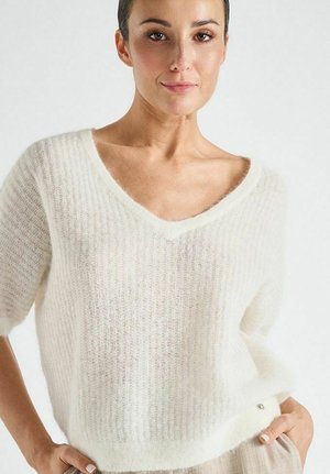 Vrouw in een zachte witte V-hals trui met korte mouwen, staand met handen in de zakken van beige broek tegen een effen lichte achtergrond.