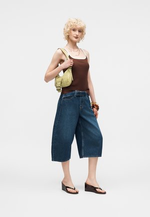 Blonde woman wearing brown tank top, wide-leg denim culottes, black wedge sandals, holding a small yellow handbag on her shoulder.