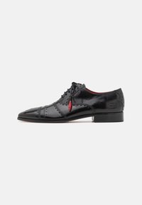 Scarpa di pelle nera con cuciture decorative, punta affusolata, chiusura con lacci e un accento rosso a contrasto vicino ai lacci.