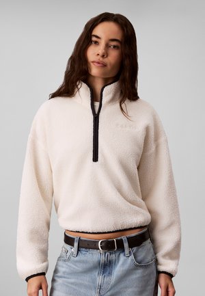 Kvinde med langt mørkt hår iført en hvid fleece-trøje med lynlås og lyseblå jeans med sort bælte mod en ensfarvet grå baggrund.