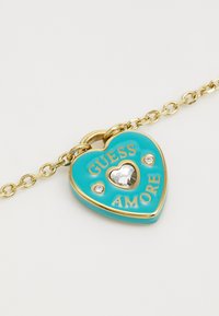 Pandantiv în formă de inimă de culoare turquoise, cu accente aurii, având textul "GUESS AMORE", o inimă oglindită și două cristale mici pe un lanț de aur.