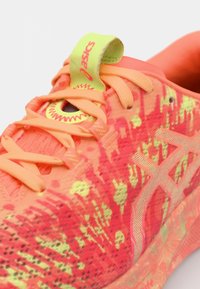 Chaussure de sport orange vif avec une tige texturée ornée de motifs verts et jaunes, d'accents réfléchissants et d'un onglet de talon vert néon.