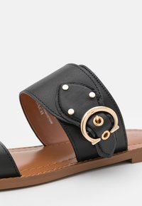 Sandale en cuir noire avec une large bride et un détail de boucle dorée sur une semelle couleur beige, vue de près sous un angle latéral.
