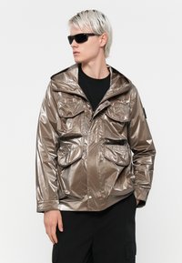 Jachetă metalică de bronz cu glugă, având patru buzunare frontale și închidere cu fermoar în față. Purtați peste un tricou negru și asortată cu ochelari de soare.