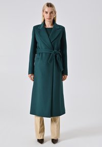 Teal wollen jas met een riem, reverskraag, zijzakken en een rechte snit, gecombineerd met beige broeken en puntige schoenen.