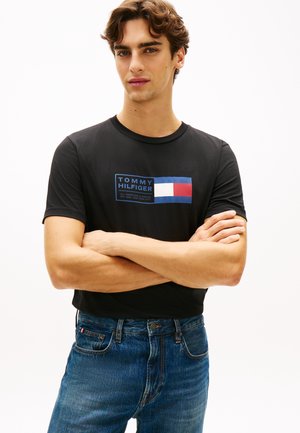 Mladi muškarac sa smeđom kosom stoji sa sklopljenim rukama, nosi crnu Tommy Hilfiger majicu i plave traperice na jednobijeloj pozadini.