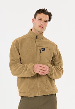 Mann trägt eine beige Fleecejacke mit Reißverschluss und Brusttasche und steht vor einem schlichten weißen Hintergrund. Die Hände sind vorne verschränkt.