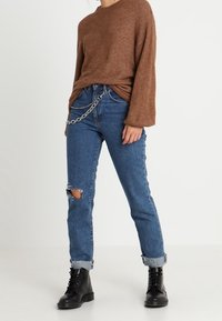 Hnědý pletený svetr, modré roztrhané džíny s ohrnutými nohavicemi, stříbrný řetězový detail. Černé taktické boty, stojící před jednobarevným pozadím.