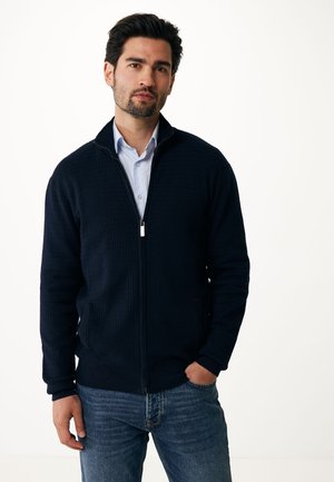 Pull zippé bleu marine avec un motif texturé, col montant, poignets côtelés et deux poches latérales, associé à une chemise bleu clair et un jean.
