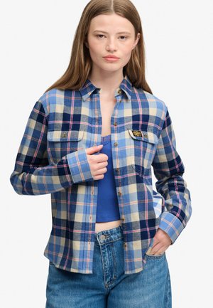 Ung kvinde med lige brunt hår iført en blå og beige ternet skjorte over en blå cropped top og blå jeans, stående med den ene hånd i lommen.
