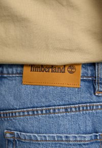 Timberland leather patch on blue denim jeans, featuring visible stitching and a tree logo. Background is a plain beige shirt fabric.