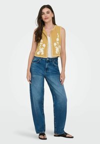 Gul ärmlös topp med vit blommig broderi, tillsammans med lösa blå denimjeans. Modellen står i svarta slidesandaler.