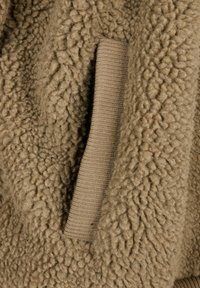 Tan jakna iz flisa s teksturirano, puhasto površino in tanjim rebrastim žepom v isti barvi. Tkanina ima mehak, udoben videz.