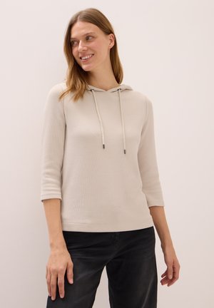 Woman smiling, looking left, wearing a beige ribbed hoodie with drawstrings and black pants against a plain light background.