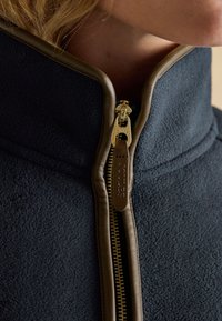 Primo piano di una giacca in pile blu navy con rifiniture in pelle marrone e una cerniera dorata parzialmente chiusa vicino al collo e al mento di una persona.