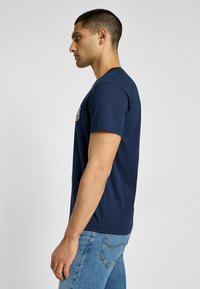 Granatowa bawełniana koszulka z krótkim rękawem z kolorowym logo na piersi. Noszona z jasnoniebieskimi dżinsami.