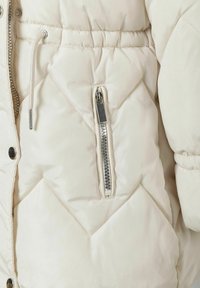 Beige Daunenjacke mit gestepptem Design, elastischem Bund und einer mit Metallakzenten versehenen, mit Reißverschluss versehenen Brusttasche sowie einem Zugband.
