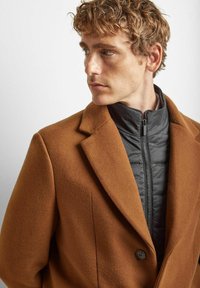 Brun ull-blend kåpe med noterte reverser og lukking med en enkelt knapp, lagd over en svart quiltet ytterjakke med glidelås.