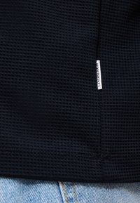 Μαύρο ύφασμα με υφή βάφλας και μια μικρή λευκή ετικέτα με την ένδειξη "LINDBERGH" κοντά στο τελείωμα πάνω από ανοιχτό μπλε τζιν.