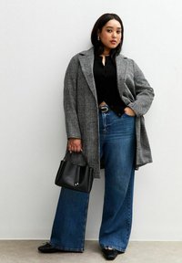 Cappotto oversize a quadri grigio, top nero, jeans a gamba larga blu e una borsa strutturata nera. Capelli sciolti, in piedi davanti a un muro chiaro.