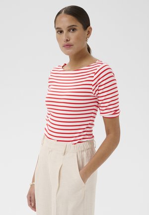 Kvinde med mørkt hår i en hestehale, iført en rød- og hvidstribet kortærmet top og beige bukser med høj talje, hånd i lommen.