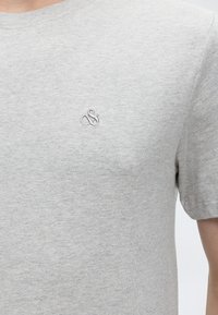 Hellgraues T-Shirt mit kurzen Ärmeln und einem kleinen bestickten Logo auf der linken Brust, getragen von einer Person, die vom Hals bis zur Mitte des Torsos gezeigt wird.