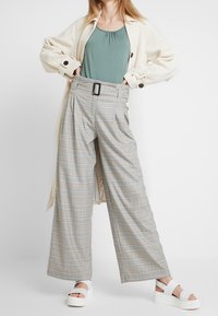 Vida byxa i ljusgrått rutigt mönster med blå detaljer, med en bältesöppning i midjan; matchas med en mintgrön tanktop och vita sandaler.