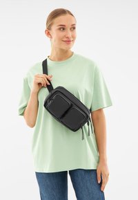 Svart nylon crossbody-väska med två fickor med dragkedja fram, hängande remmar och en slät textur, bärs över en ljusgrön t-shirt.