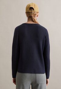 Marineblauer gestrickter Pullover mit langen Ärmeln und rundem Ausschnitt, kombiniert mit hellgrauen, maßgeschneiderten Hosen; glatte Textur und figurbetontes Design.