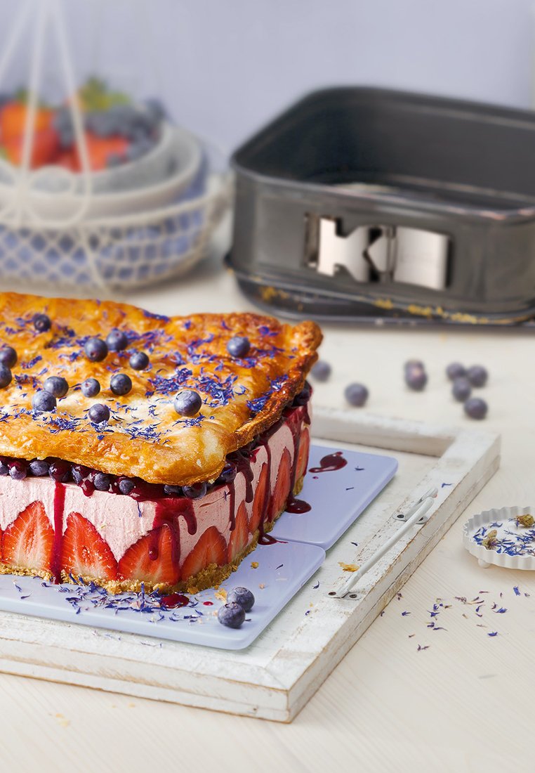 Lagerad dessert med en gyllenbrun skorpa, garnerad med blåbär och lavendel, på en vit träbricka med blå tallrikar.