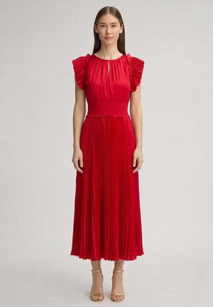 Noor naine seisab, seljas voldiline punane midi-kleit, rüüsidega lühikesed varrukad ja kuldsed pahkluu-ümber rihmadega kontsakingad ühtlasel taustal.