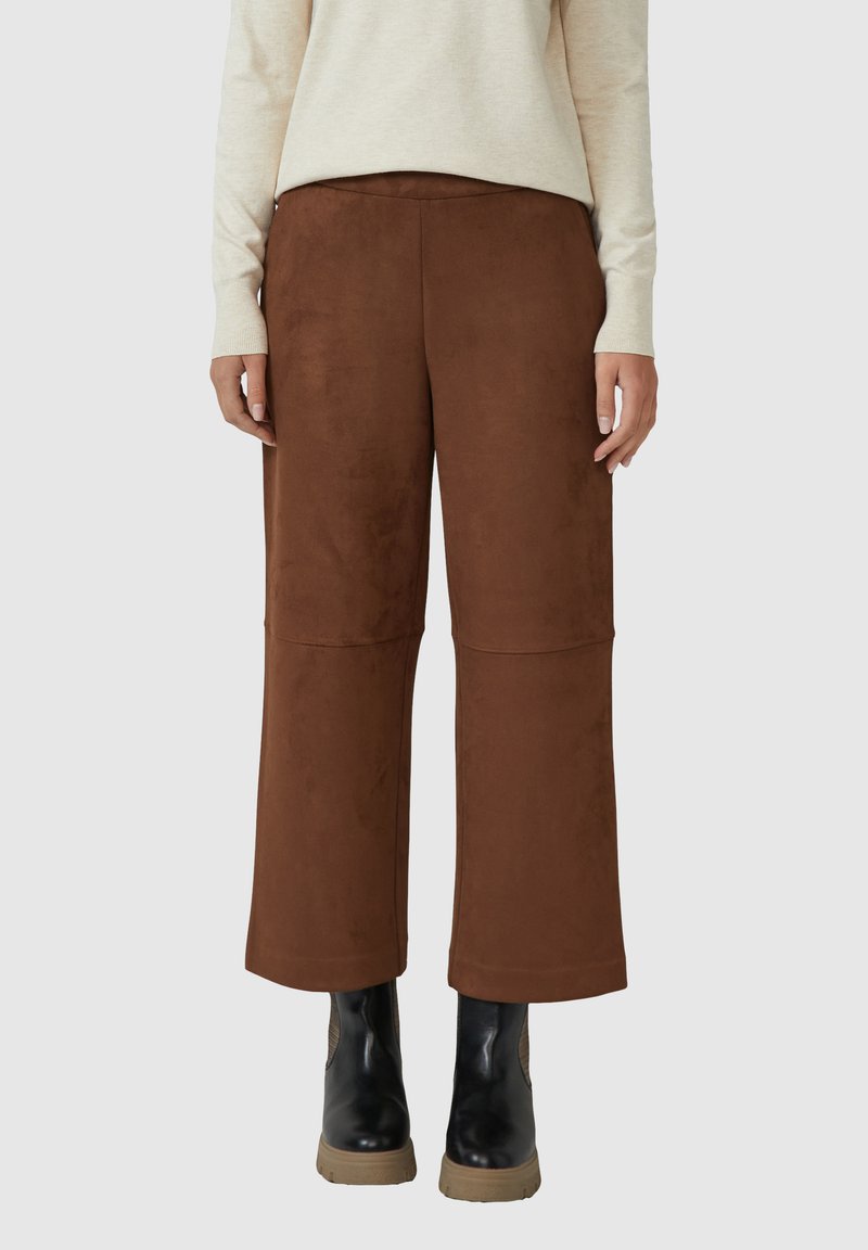 Braune Wildleder-Hosen mit weitem Bein, die eine glatte Textur und Nahtdetails aufweisen, kombiniert mit schwarzen Knöchelstiefeln mit einer cognacfarbenen Sohle.