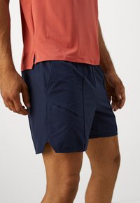 Navy athletic shorts with a textured finish, featuring a side zip pocket and slight side slits for flexibility. Paired with a coral shirt.