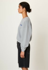 Felpa grigia chiara con collo a giro, spalle basse e ricamo blu "American Vintage", abbinata a una gonna di jeans nera.