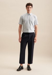 Lichtblauw en wit geruit shirt met korte mouwen, gedragen met donkere marineblauwe broek. Model draagt zwarte schoenen en staat op een neutrale achtergrond.