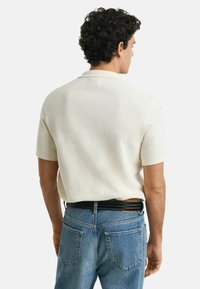 Gestricktes Poloshirt mit kurzen Ärmeln in Creme, das ein strukturiertes Muster aufweist, kombiniert mit denimblauen Jeans und einem sichtbaren schwarzen Gürtel.