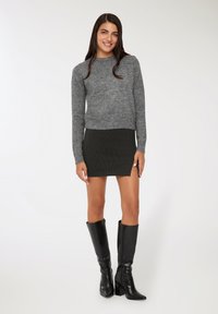 Donna sorridente, che indossa un maglione grigio a maglia, una mini gonna nera a righe con spacco laterale e stivali alti al ginocchio neri con tacco, in piedi su uno sfondo bianco.