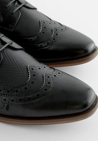 Mustad mustad nahast kleitkingad brogue detailidega, tekstuuriga ülemine osa, terav nina ja pruun kummitald. Omab lipsukujundust ja perforeeritud aktsente.