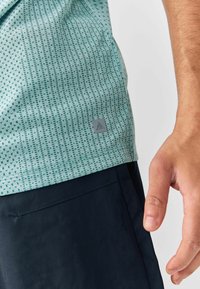 Világos türkizkék mintás ing, kicsi geometrikus motívumokkal; az anyag textúrált hatású. Szürke márkacímke az alsó bal részen. Sötét szoknya látható az alsó részen.