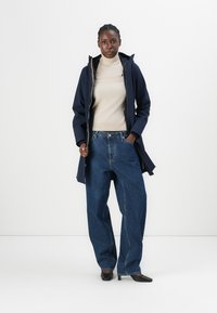 Manteau imperméable marine avec fermeture éclair, pull col roulé beige, jean bleu ample et chaussures noires à bout pointu, sur fond uni.